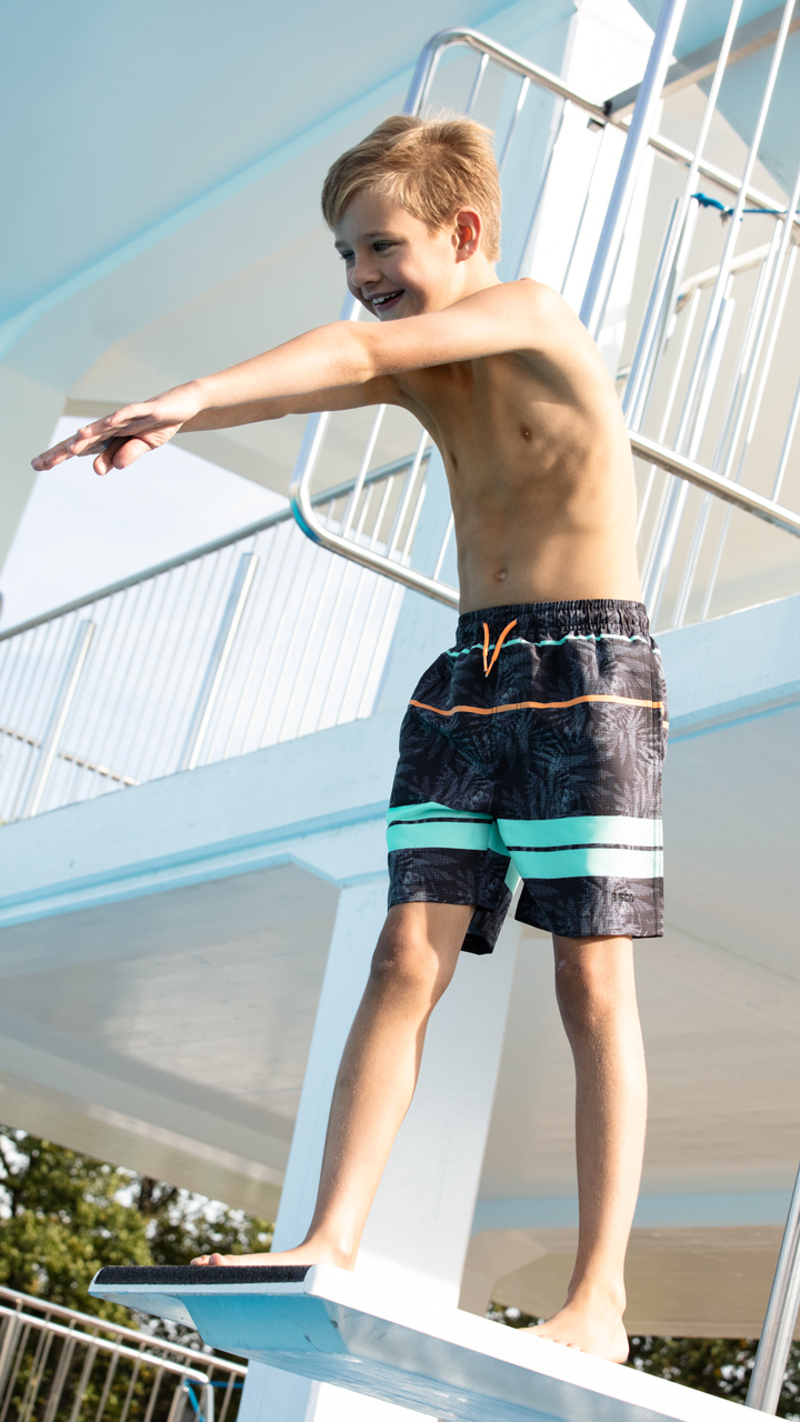 Photo of a young person in black patterned swimming shorts standing on a diving board.